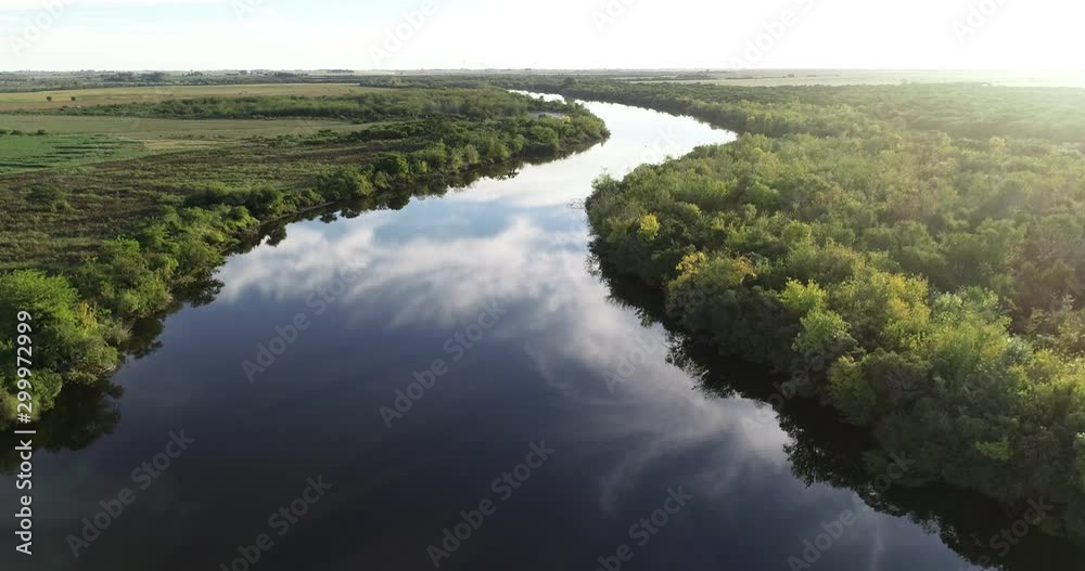Aerial of river with natural vegetation at riverbends and reflection of sky over water surface. Ascending discovering agricultural landscape and sunset at background. Dolores, Soriano, Uruguay Aerial of river with natural vegetation at riverbends and reflection of sky over water surface. Ascending discovering agricultural landscape and sunset at background. Dolores, Soriano, Uruguay