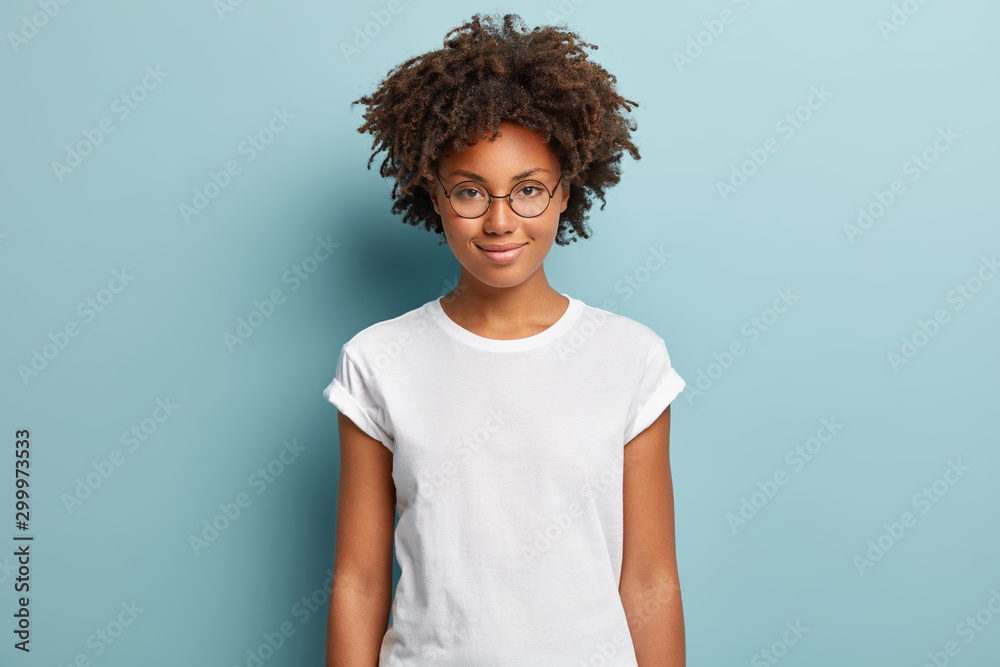 Attractive female student with curly hair, wears transparent glasses, white t shirt, stands against blue background, has calm face expression, tender smile, listens interlocutor with pleasure - obrazy, fototapety, plakaty 