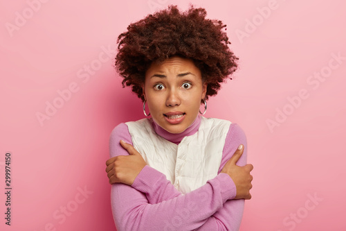 Shot of scared young girl crosses hands over chest, trembles and looks puzzled at camera, feels cold and chill, tries to protect herslelf wears not enough warm clothes, isolated on pink wall