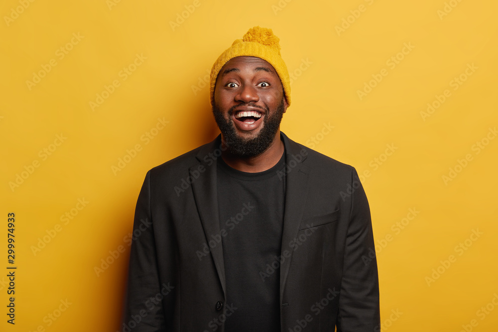 Shot of glad plump dark skinned man laughs positively, smiles joyfully, rejoices pleasant meeting, wears black elegant clothes and bright headgear, stands against yellow background, feels happiness Shot of glad plump dark skinned man laughs positively, smiles joyfully, rejoices pleasant meeting, wears black elegant clothes and bright headgear, stands against yellow background, feels happiness