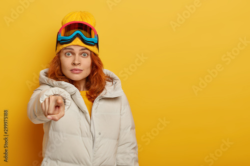 Impressed ginger woman snowboarder points into camera, indicates at ski slope, dressed in outerwear, protective snowboarding goggles, isolated over yellow background. Ski winter resort concept