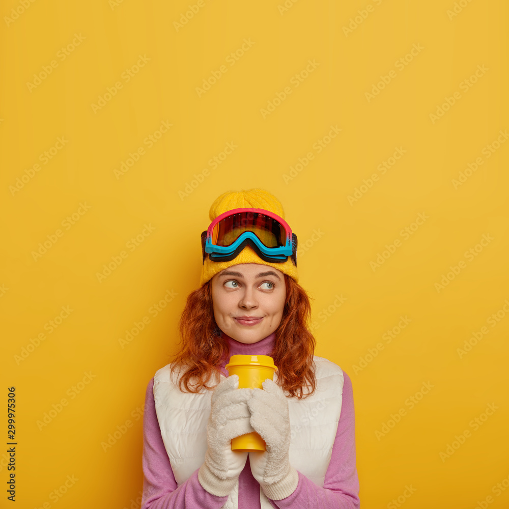 Winter holiday concept. Attractive ginger girl has happy thoughtful expression, holds takeout coffee, drinks hot beverage, visits winter park resort, relaxes and warms up during frosty cold weather Winter holiday concept. Attractive ginger girl has happy thoughtful expression, holds takeout coffee, drinks hot beverage, visits winter park resort, relaxes and warms up during frosty cold weather