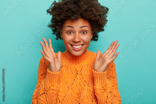 Close up shot of joyful curly woman expresses enthusiasm and joy, keeps palms raised, smiles broadly, wears bright winter sweater, enjoys amusing night with friends, isolated on blue background