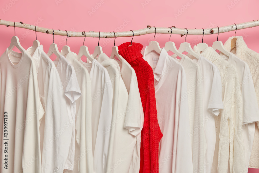 Choice of white clothes on hangers in fashion store. Red bright knitted sweater between outfits in one tone, isolated on pink wall. Fashion background. Several jumpers and tshirts on racks. Sell out