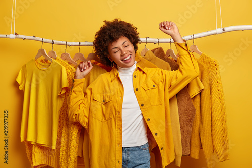 Carefree curly woman clothes buyer dances with happiness, raises arms, buys yellow clothing from new collection, rejoicing successful shopping day, being in high spirit, dances against outfits on rack