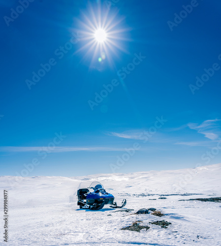 One blue snowmobile stands on a crust snow in Norwegian mountains, warm day and very bright sun, no clouds, snow starting to smelt, snowmobile road, very far away leads to the tops of mountains