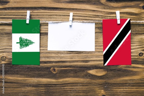 Hanging flags of Norfolk Island and Trinidad And Tobago attached to rope with clothes pins with copy space on white note paper on wooden background.Diplomatic relations between countries. photo