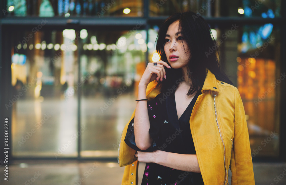 Fototapeta premium Attractive asian female with long dark hair looking pensively aside while standing on an urban background. Hipster girl waiting for the friends next to the shopping mall entrance to spend spare time