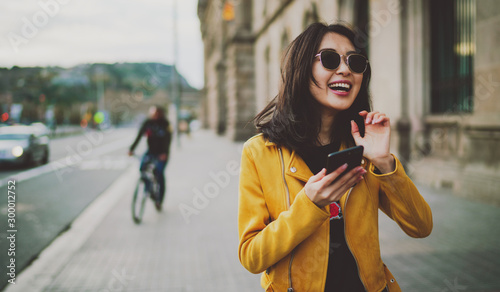 Cheerful asian student girl wearing modern sunglasses laughing at friends' photos in social media by a mobile phone. Happy model look woman in casual outfit checking blog comments via smartphone. photo