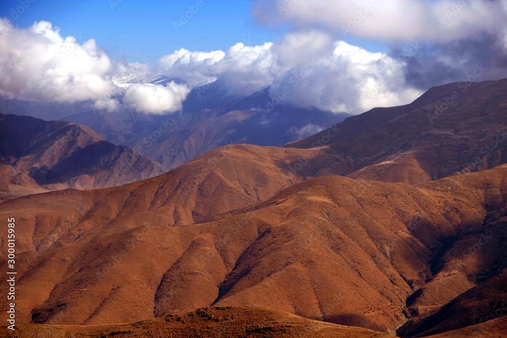 Panoramic view of the Tibetan Plateau showing brown mountains against a blue sky covered by white clouds.