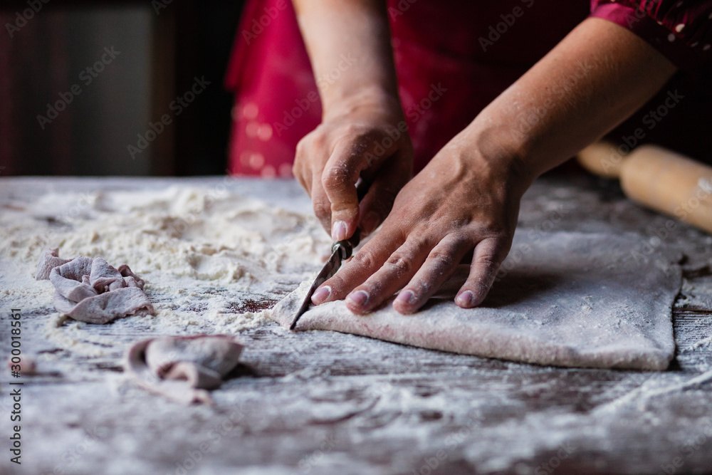 Woman in red dress and apron is preparing a dough for delicious homemade pink pasta on her kitchen. Dark and moody, low key. Flour all over