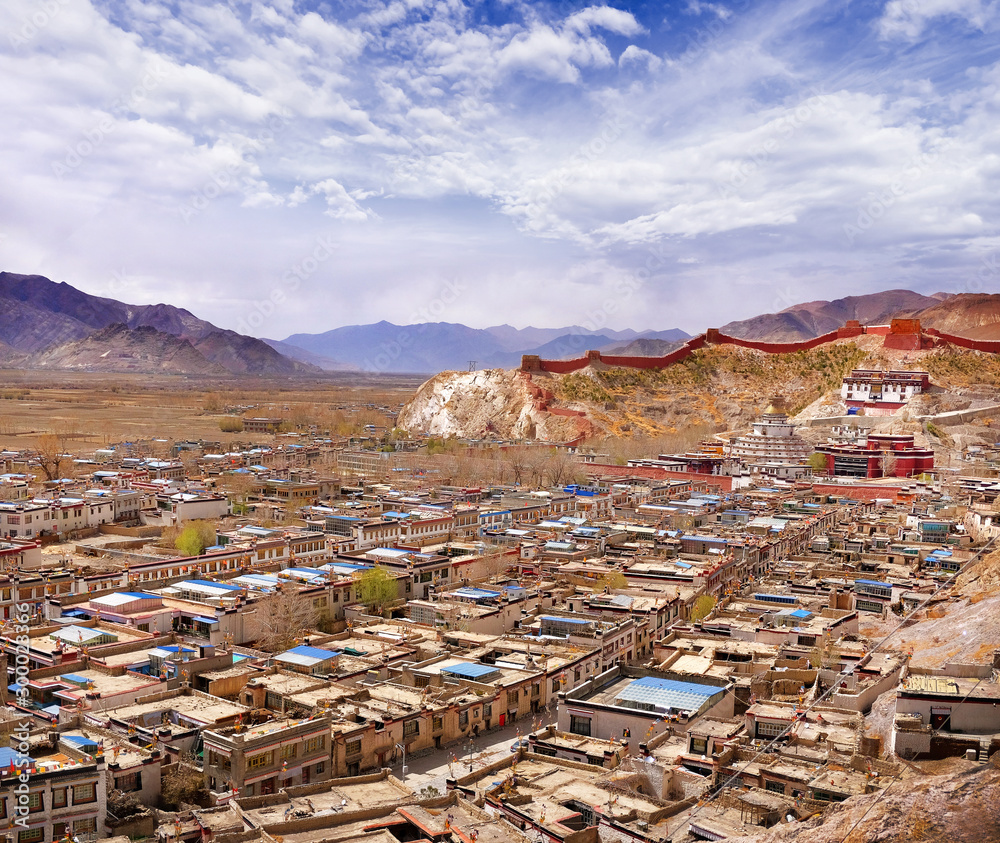 View of traditional Tibetan village surrounded by the Himalayan Mountains, against a blue sky covered by white clouds.