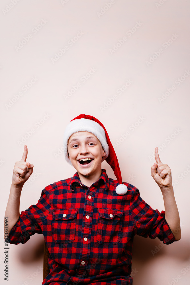 Funny boy in santa hat pointing upwards with index fingers over empty background. Advertisement, banner or poster template, Christmas, winter holidays preparation Funny boy in santa hat pointing upwards with index fingers over empty background. Advertisement, banner or poster template, Christmas, winter holidays preparation