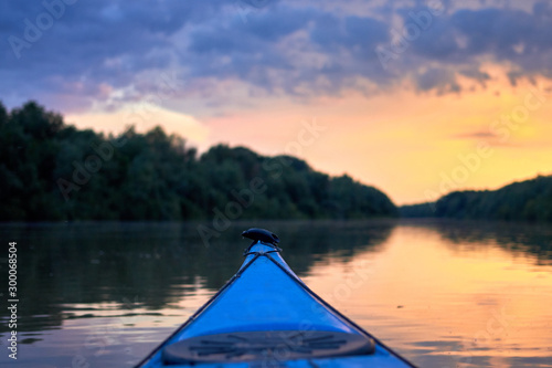 Bow of blue kayak. Dramatic sunset over Danube river with forest along riverside