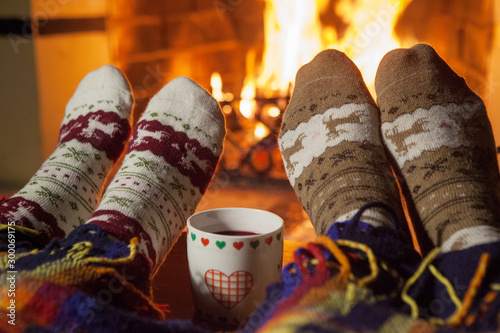 Man and woman in warm knitted socks with cups of hot punch in front of the fireplace
