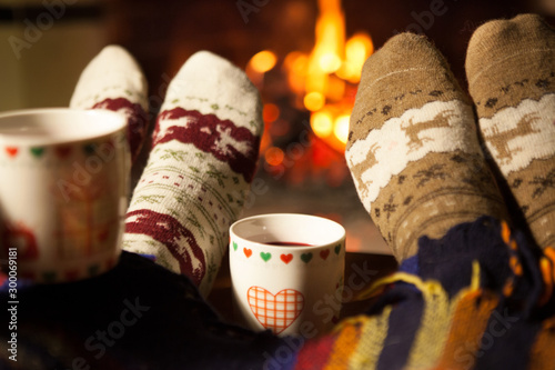 Man and woman in warm knitted socks with cups of hot punch in front of the fireplace