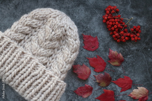 White winter handmade knitted hat with rowan berries and red autumn maple leaves on grey marble background  vertical