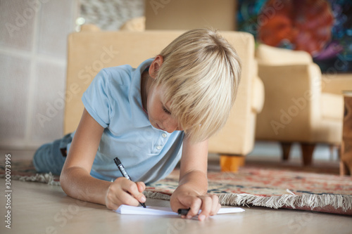 a teenager blonde boy lies on the floor in the room and writes on a paper airplane.