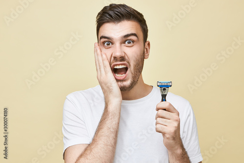 Portrait of frustrated unhappy young European man with stubble holding hand on his cheek and screaming, having terriified look, suffering from skin irritation because of shaving, holding razor stick