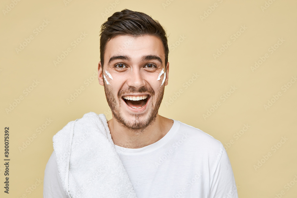 Obraz premium Studio shot of emotional excited young bearded guy posing isolated in t-shirt and towel on his shoulder going to shave and take shower after training in gym, stripes of white foam on his cheeks