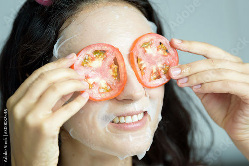 A beautiful woman wearing a towel and a white bathrobe has mask on face-covering eyes with sliced tomatoes with happy and relaxing on the bed at a condominium in the morning.
