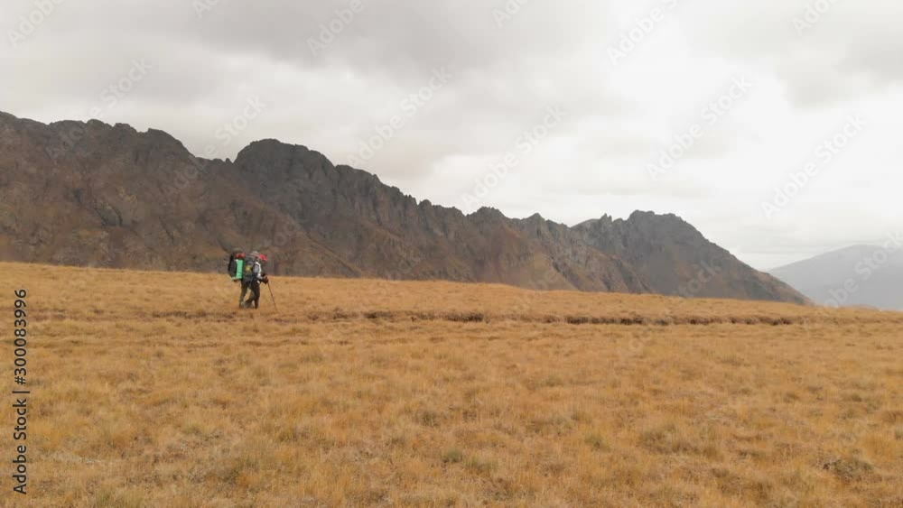 Aerial view of a couple of travelers man and woman with large backpacks in hats and sunglasses walk along an alpine plateau surrounded by epic mountains Aerial view of a couple of travelers man and woman with large backpacks in hats and sunglasses walk along an alpine plateau surrounded by epic mountains