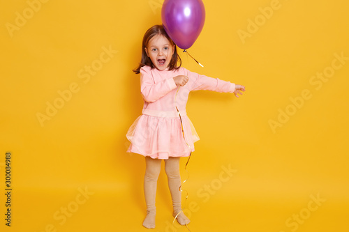 Horizontal shot of little girl with purple balloon isolated over yellow studio background  female child shouting somethig  celebrating herbirtday  kid wearing rosy dress and having dark hair.