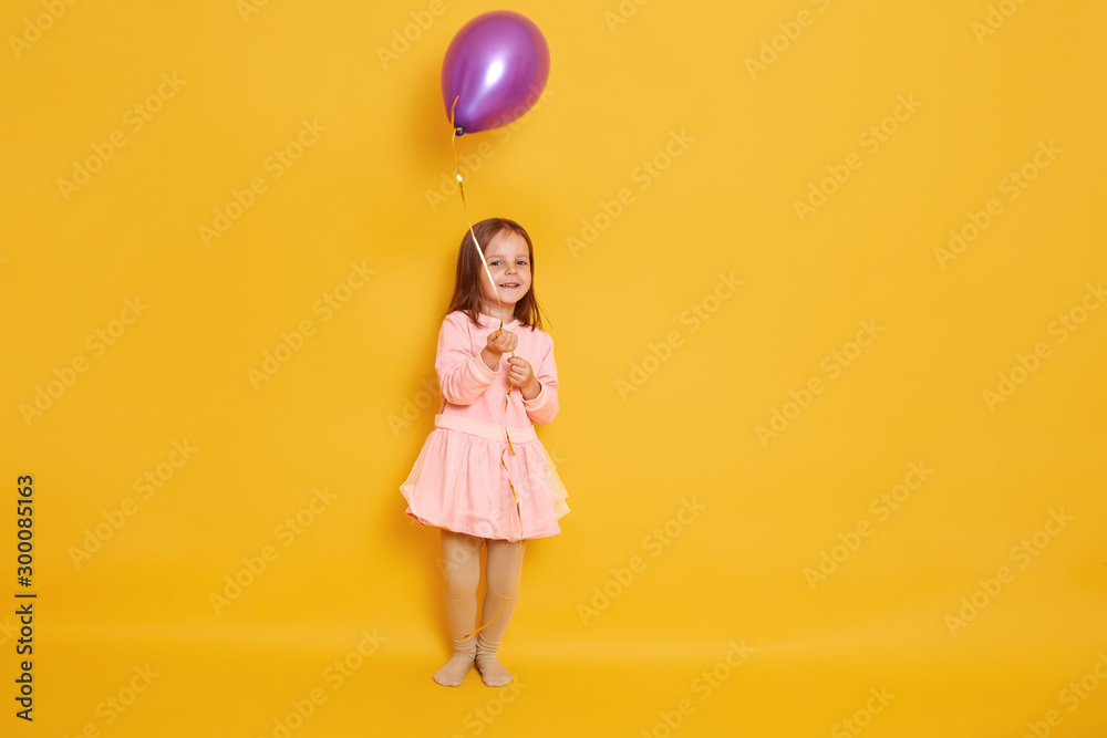 Studio shot of little girl wearing rose dress playing with purple balloon isolated over yellow background, having birthday, having fun with balloon, cute female child with dark hair. Childhood concept - obrazy, fototapety, plakaty 