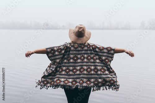 Tela Woman in hat and poncho standing on the river bank and looking towards autumn fo