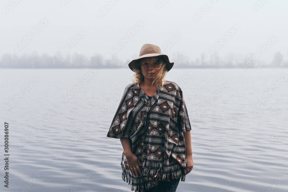 Woman in hat and poncho standing on the river bank and looking towards autumn forest covered with morning fog