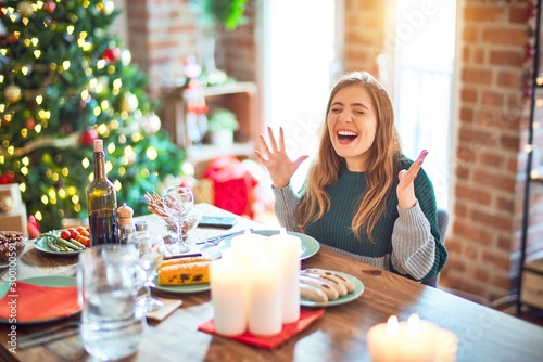 Slika na platnu Young beautiful woman sitting eating food around christmas tree at home celebrating mad and crazy for success with arms raised and closed eyes screaming excited