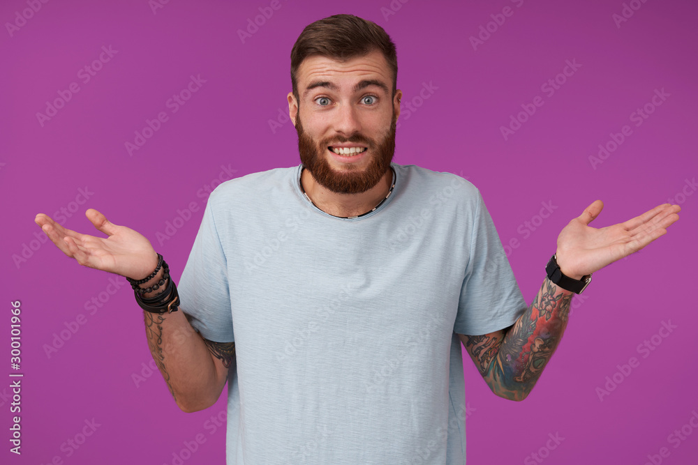 Portrait of young unshaved tattooed brunette man wearing blue t-shirt and trendy accessories contracting forehead and shrugging with raised palms, isolated over purple background Portrait of young unshaved tattooed brunette man wearing blue t-shirt and trendy accessories contracting forehead and shrugging with raised palms, isolated over purple background