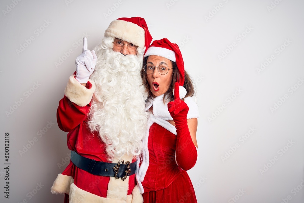 Middle age couple wearing Santa costume hugging over isolated white background pointing finger up with successful idea. Exited and happy. Number one. Middle age couple wearing Santa costume hugging over isolated white background pointing finger up with successful idea. Exited and happy. Number one.
