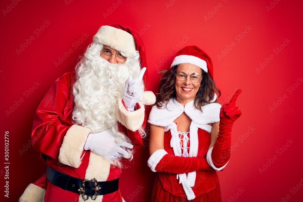 Middle age couple wearing Santa costume and glasses over isolated red background with a big smile on face, pointing with hand finger to the side looking at the camera. Middle age couple wearing Santa costume and glasses over isolated red background with a big smile on face, pointing with hand finger to the side looking at the camera.