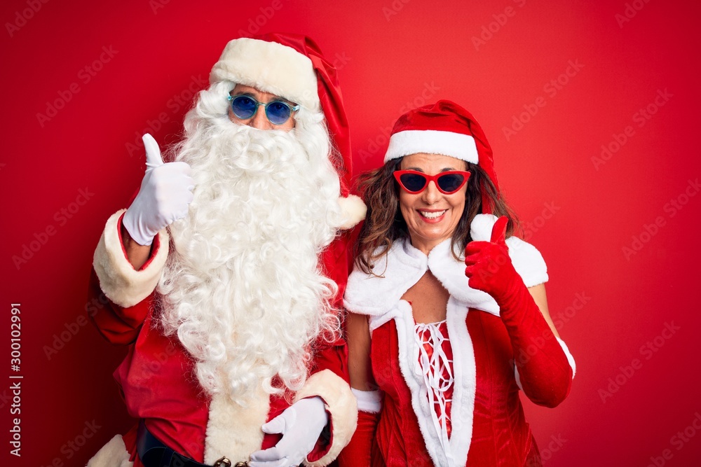 Middle age couple wearing Santa costume and sunglasses over isolated red background doing happy thumbs up gesture with hand. Approving expression looking at the camera showing success. Middle age couple wearing Santa costume and sunglasses over isolated red background doing happy thumbs up gesture with hand. Approving expression looking at the camera showing success.