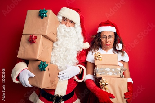 Middle age couple wearing Santa costume holding tower of gifts over isolated red background depressed and worry for distress, crying angry and afraid. Sad expression.