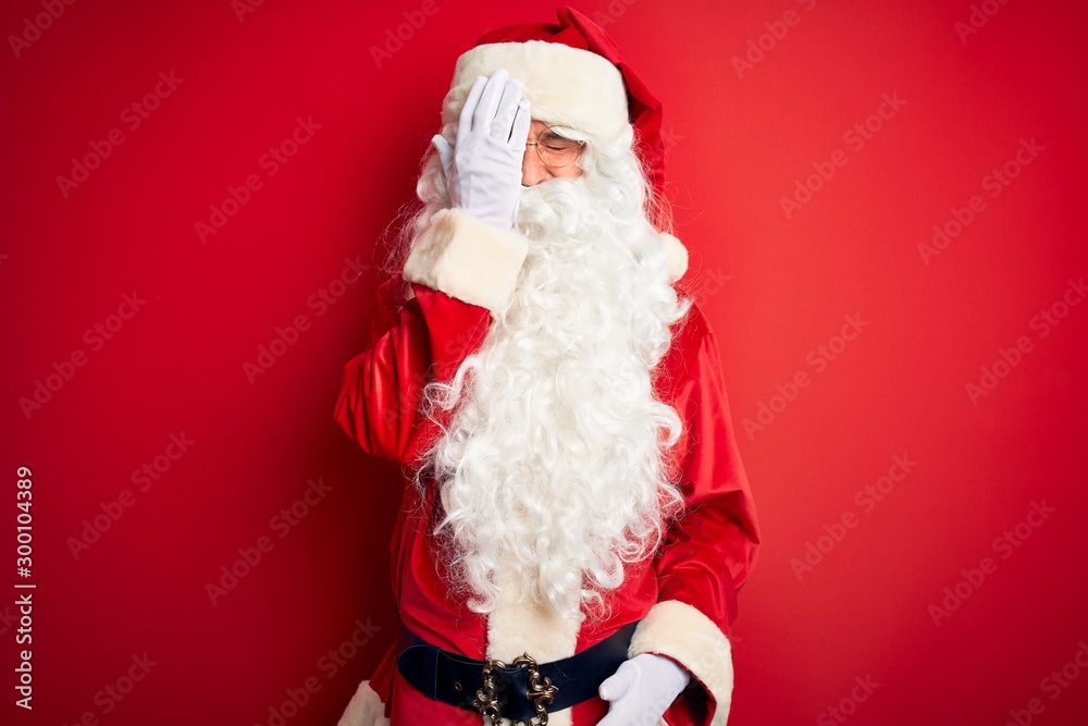 Middle age handsome man wearing Santa costume standing over isolated red background covering one eye with hand, confident smile on face and surprise emotion.