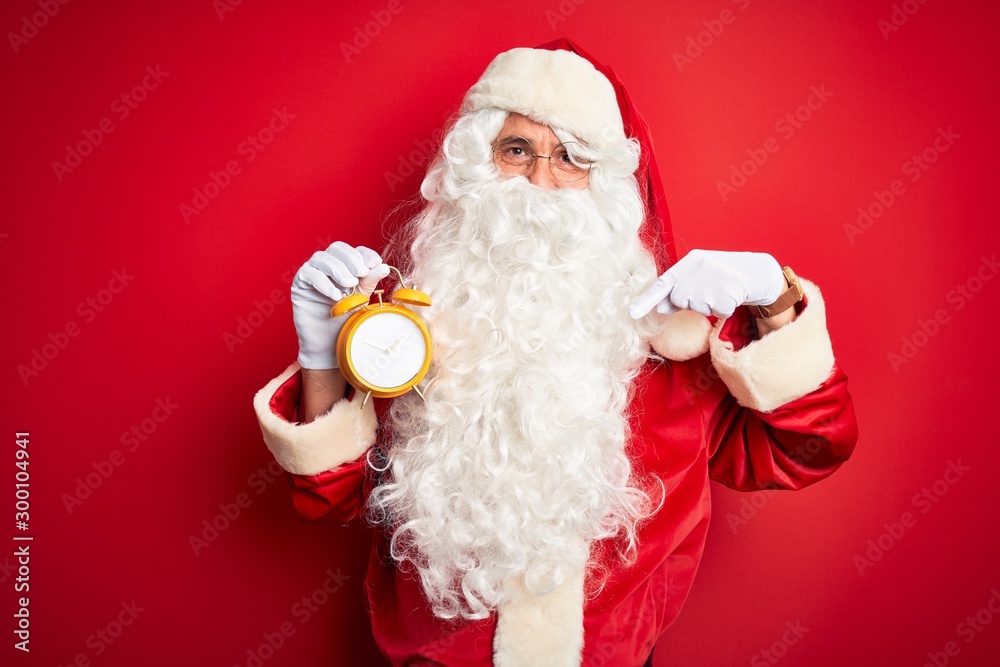 Senior man wearing Santa Claus costume holding alarm clock over isolated red background with surprise face pointing finger to himself
