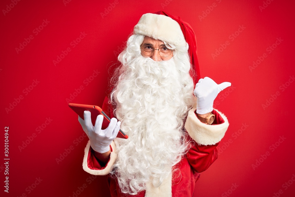 Middle age man wearing Santa Claus costume using tablet over isolated red background pointing and showing with thumb up to the side with happy face smiling Middle age man wearing Santa Claus costume using tablet over isolated red background pointing and showing with thumb up to the side with happy face smiling