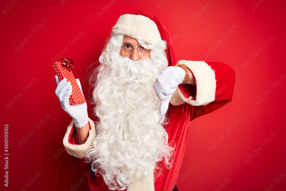 Middle age man wearing Santa Claus costume holding gift over isolated red background with angry face, negative sign showing dislike with thumbs down, rejection concept