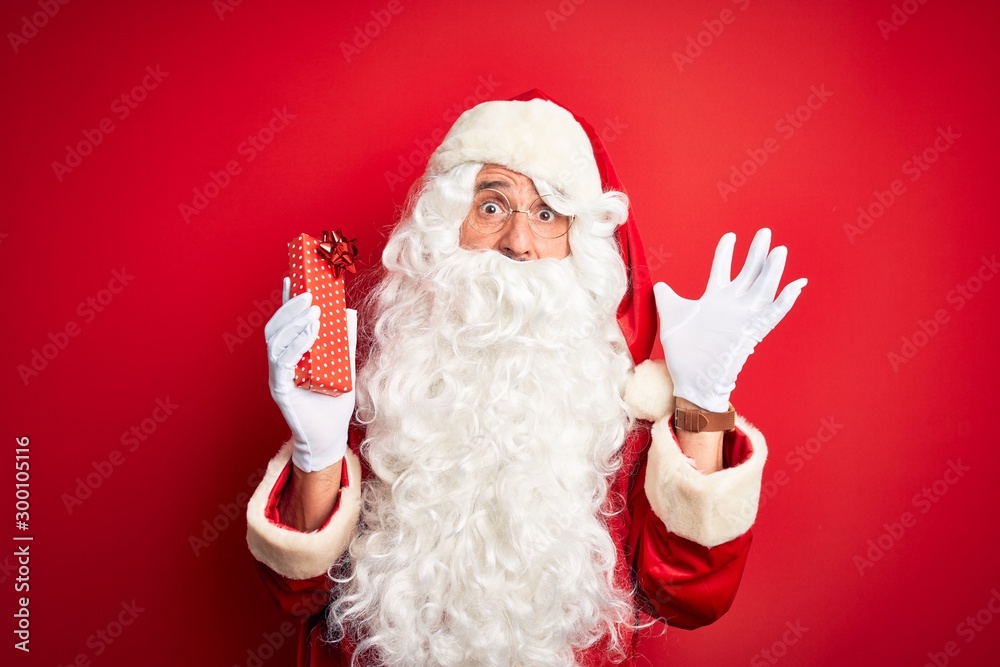 Middle age man wearing Santa Claus costume holding gift over isolated red background very happy and excited, winner expression celebrating victory screaming with big smile and raised hands Middle age man wearing Santa Claus costume holding gift over isolated red background very happy and excited, winner expression celebrating victory screaming with big smile and raised hands
