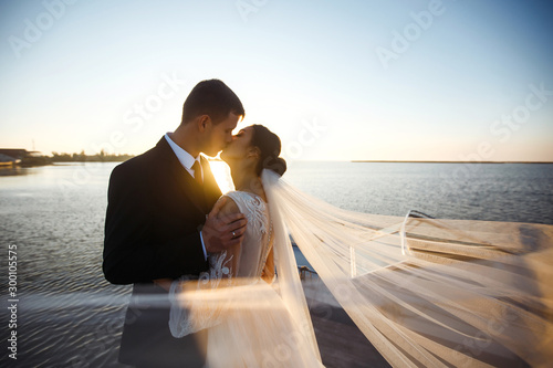 Pretty bride and stylish groom together on the bridge against the background of the boat. Newlyweds enjoy each other tenderly in the shadow of a flying veil. Together. Wedding. Love.