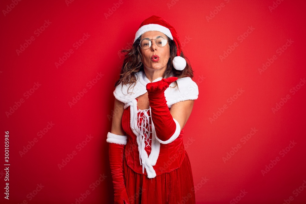 Middle age beautiful woman wearing Santa Claus costume over isolated red background looking at the camera blowing a kiss with hand on air being lovely and sexy. Love expression. Middle age beautiful woman wearing Santa Claus costume over isolated red background looking at the camera blowing a kiss with hand on air being lovely and sexy. Love expression.