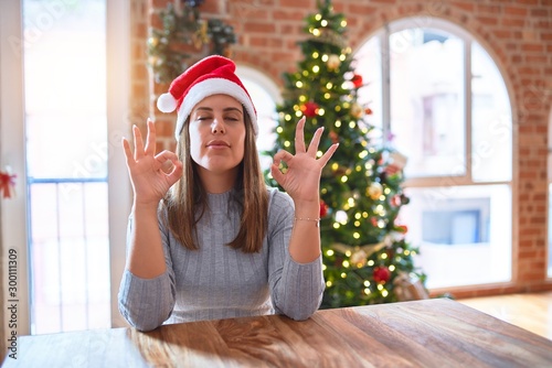 Young beautiful woman wearing santa claus hat at the table at home around christmas decoration relax and smiling with eyes closed doing meditation gesture with fingers. Yoga concept.