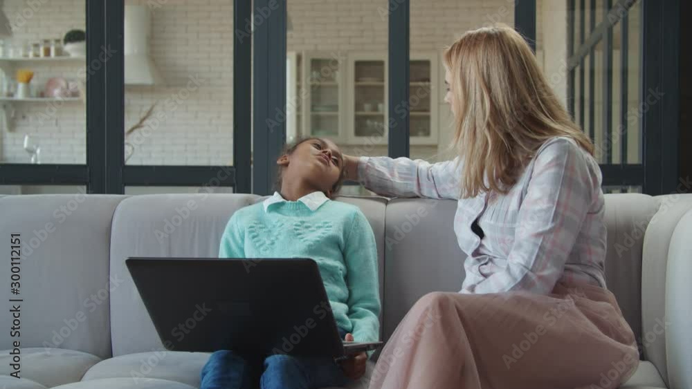 Worried caucasian mother checking temprerature of sick mixed race daughter by hand, touching her forehead while sitting on couch in domestic room. Caring mon hand taking temperature of ill child.