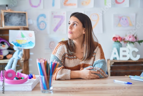 Young beautiful teacher woman wearing sweater and glasses sitting on desk at kindergarten looking to side, relax profile pose with natural face with confident smile.