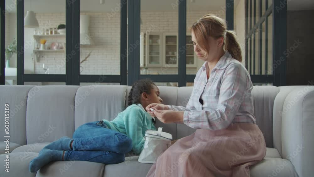 Caring affectionate caucasian mother checking temperature of ill mixed race girl with digital thermometer at home. Sick sad daughter lying on sofa and worried mom measuring temprature indoors. Caring affectionate caucasian mother checking temperature of ill mixed race girl with digital thermometer at home. Sick sad daughter lying on sofa and worried mom measuring temprature indoors.