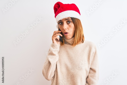 Beautiful redhead woman wearing christmas hat talking on smartphone scared in shock with a surprise face  afraid and excited with fear expression
