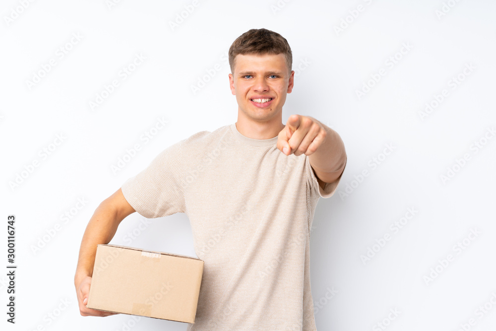 Young handsome man over isolated white background holding a box to move it to another site while pointing to the front Young handsome man over isolated white background holding a box to move it to another site while pointing to the front