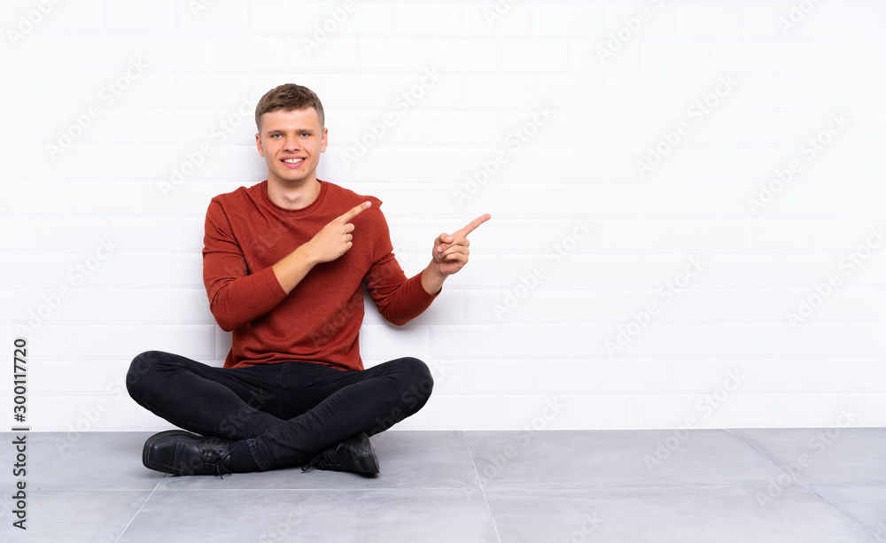 Young handsome man sitting on the floor pointing finger to the side